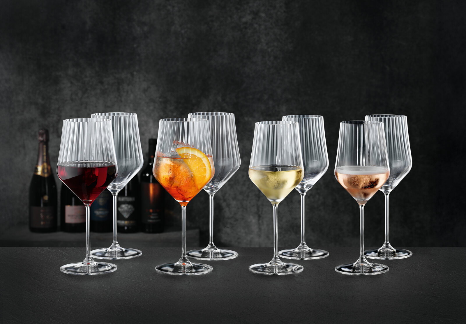 Mehrere geriffelte Weingläser mit Stiel auf dunklem Untergrund, teils mit Rotwein, Aperol-Spritz, Weißwein und Rosé gefüllt, frontal in leichter Seitenperspektive fotografiert.