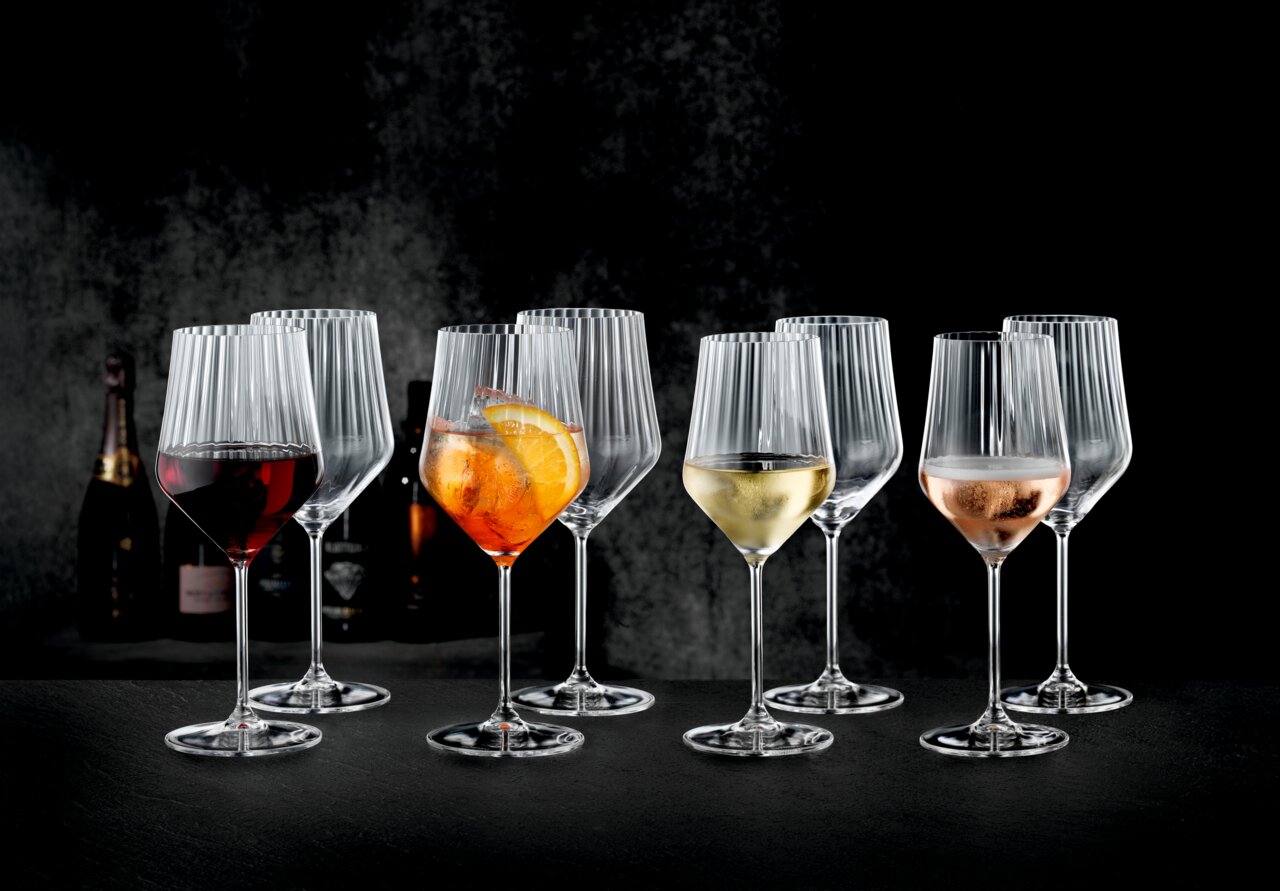 Mehrere geriffelte Weingläser mit Stiel auf dunklem Untergrund, teils mit Rotwein, Aperol-Spritz, Weißwein und Rosé gefüllt, frontal in leichter Seitenperspektive fotografiert.