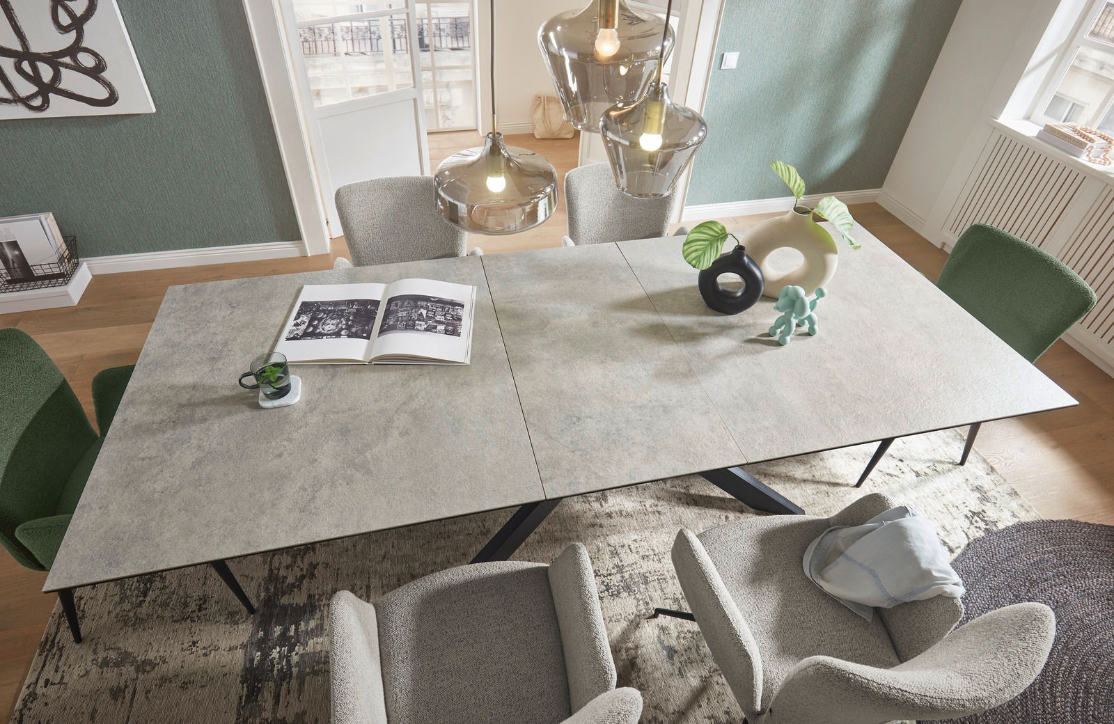 Moderner Esstisch mit grauer Steinoptik-Oberfläche, umgeben von grünen und grauen Stühlen, aus der Vogelperspektive fotografiert. Dekoriert mit Vasen, einer Pflanze und einem offenen Buch.
