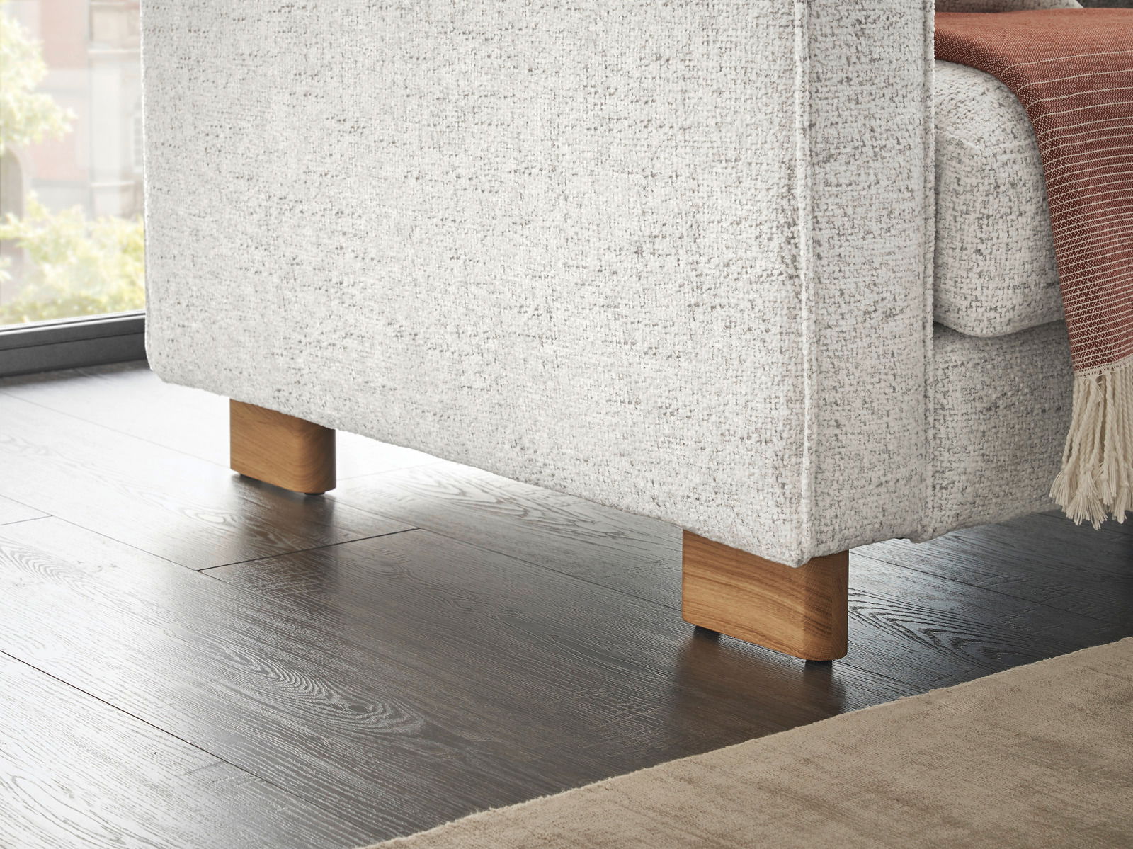 Nahaufnahme der unteren Ecke eines Ecksofas mit hellgrauem Stoffbezug und Holzfüßen, Perspektive von der Seite.