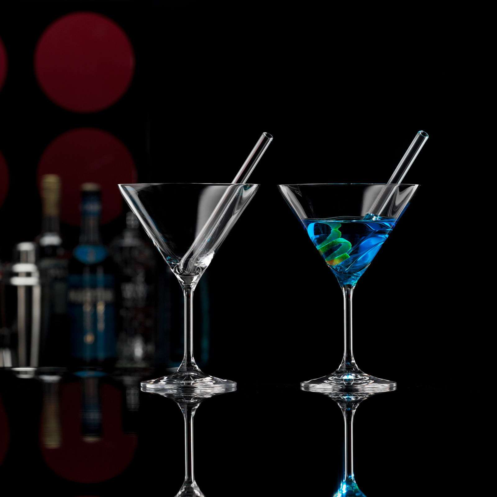 Novel Cocktailschale 2er-Set mit Trinkhalm Zwei Cocktailschalen mit Trinkhalmen, eine mit blauem Getränk und Zitronenspirale, auf schwarzem Hintergrund mit Barzubehör im Hintergrund, seitliche Perspektive.