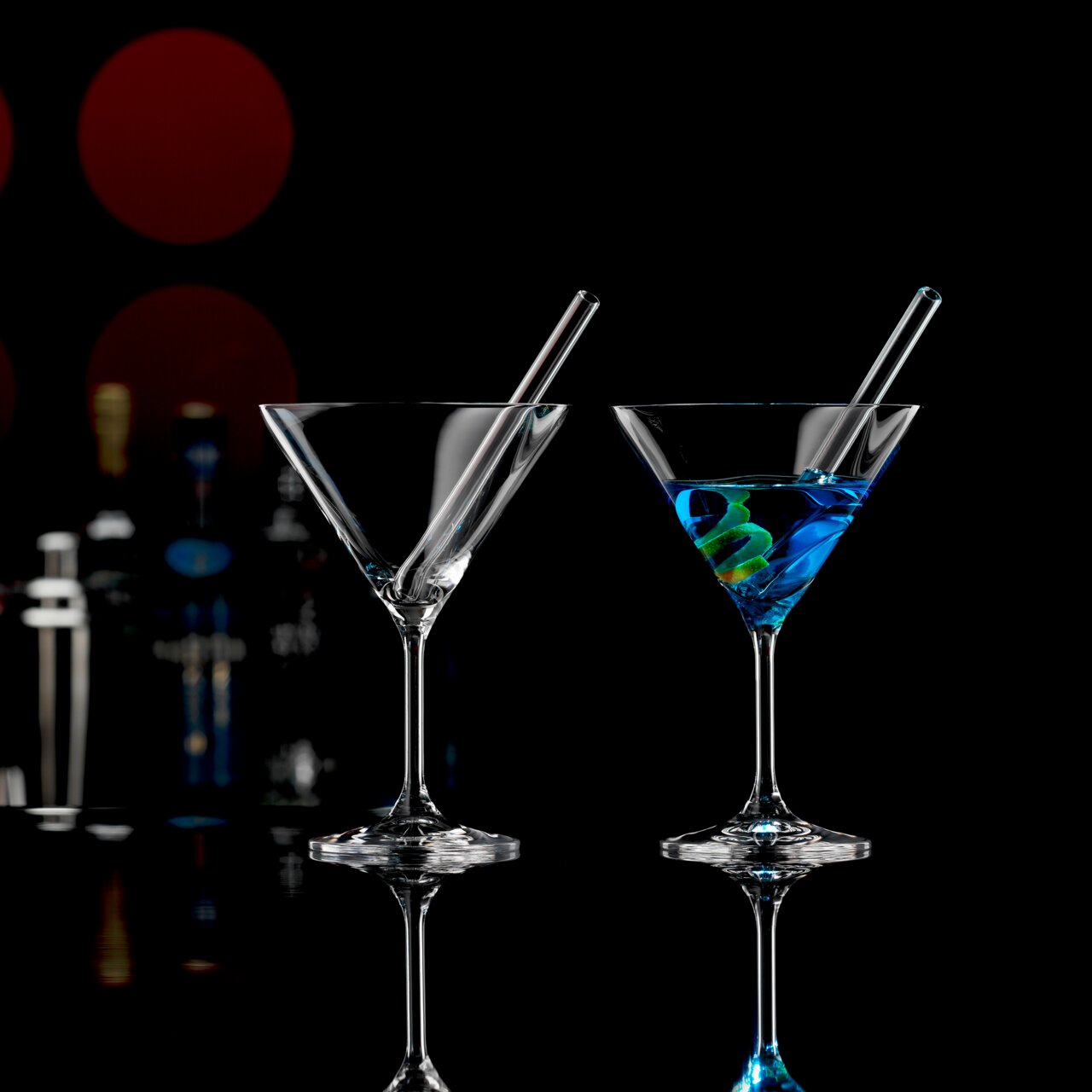 Zwei Cocktailschalen mit Trinkhalmen, eine mit blauem Getränk und Zitronenspirale, auf schwarzem Hintergrund mit Barzubehör im Hintergrund, seitliche Perspektive.