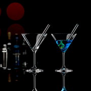Zwei Cocktailschalen mit Trinkhalmen, eine mit blauem Getränk und Zitronenspirale, auf schwarzem Hintergrund mit Barzubehör im Hintergrund, seitliche Perspektive.