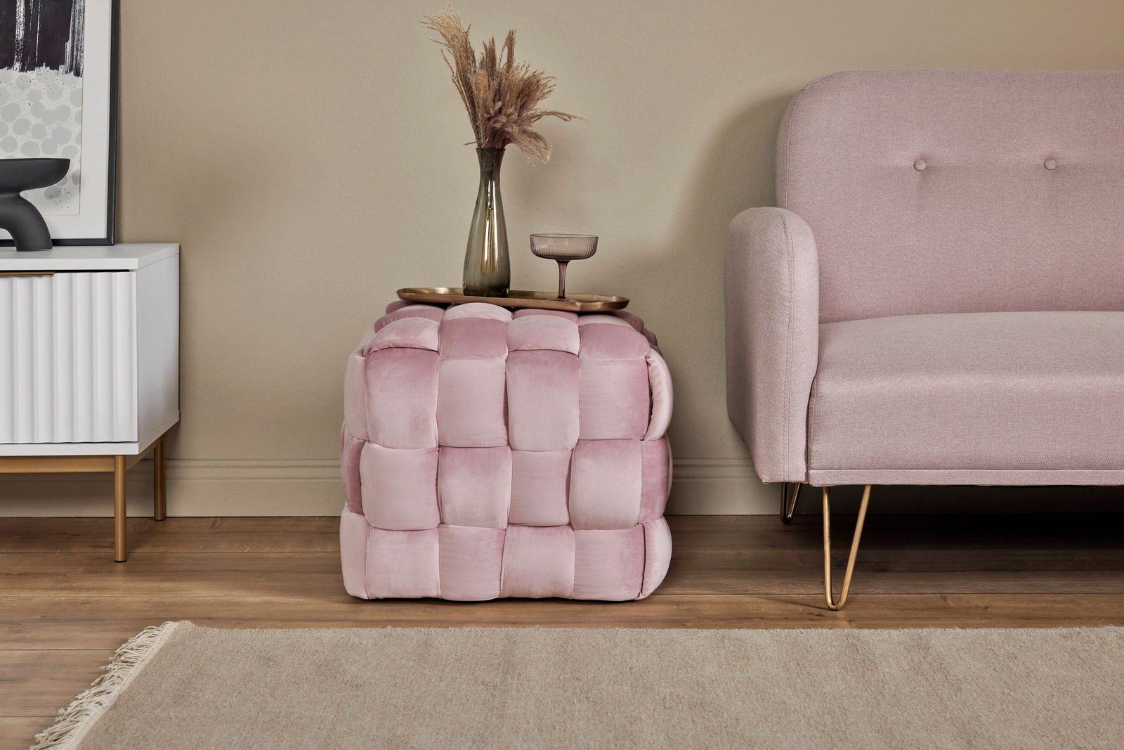 Rosa gepolsterter Hocker in Würfelform, seitlich neben einem Sofa platziert, mit einer Vase und einem Glas darauf.