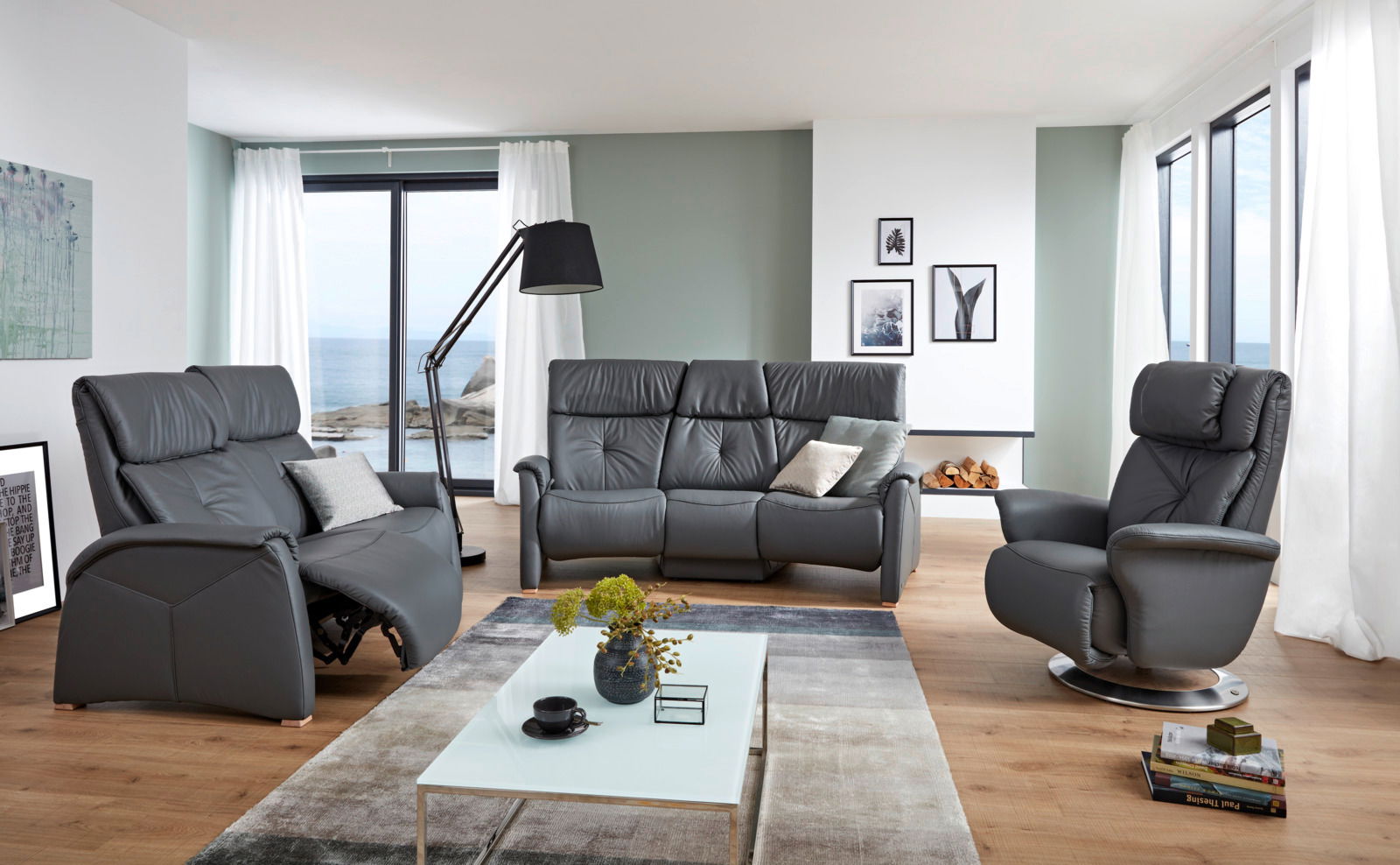 Modernes Wohnzimmer mit einem grauen 3er-Sofa in der Mitte, flankiert von einem Sessel und einem Relaxsessel, mit Blick auf das Meer durch große Fenster.