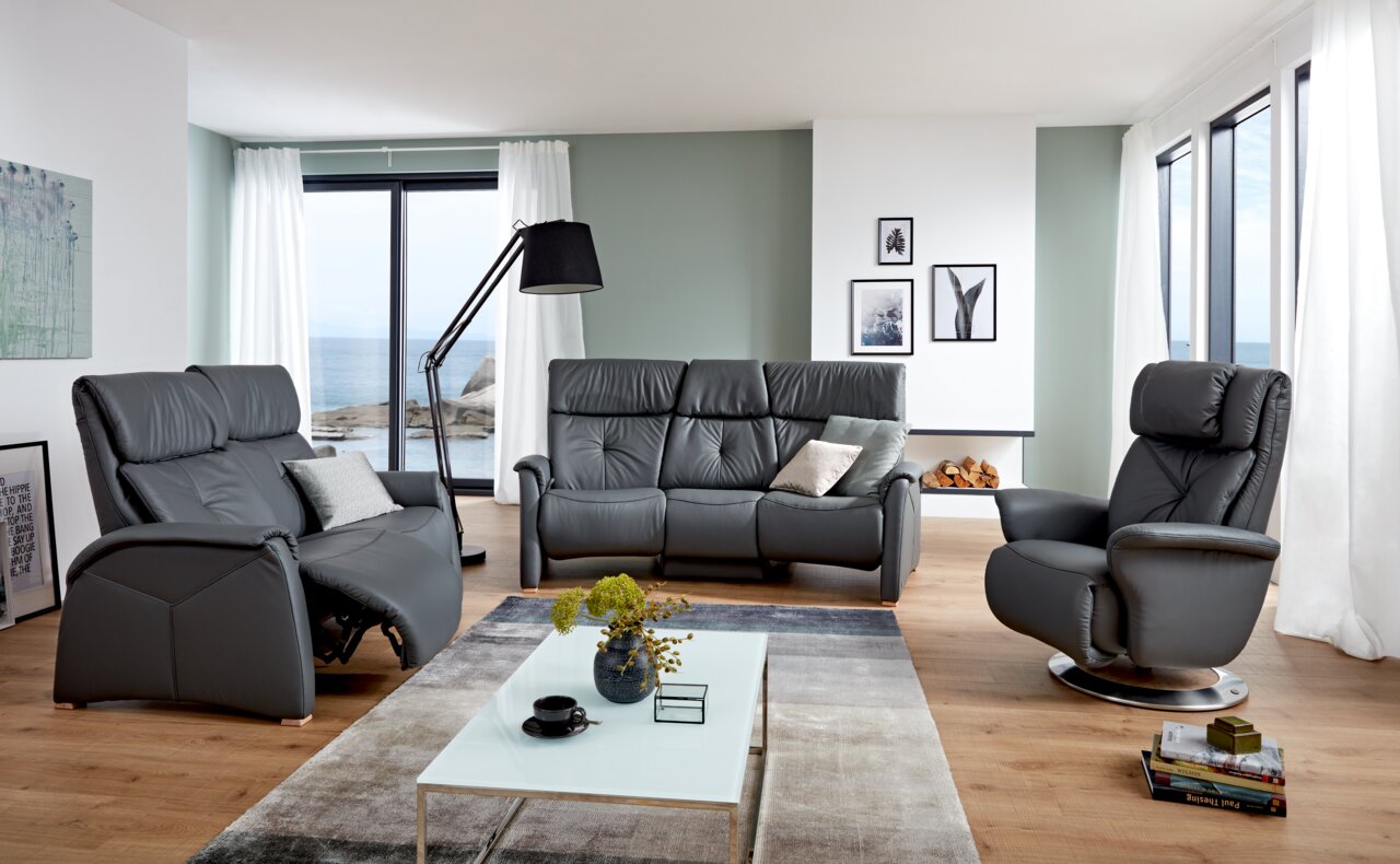 Modernes Wohnzimmer mit einem grauen 3er-Sofa in der Mitte, flankiert von einem Sessel und einem Relaxsessel, mit Blick auf das Meer durch große Fenster.