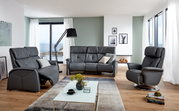 Moderne woonkamer met een grijze driezitsbank in het midden, geflankeerd door een fauteuil en een relaxstoel, met uitzicht op zee door grote ramen.