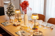Festlich gedeckter Holzesstisch in schräger Seitenansicht mit zwei warm leuchtenden Tischlampen, Kristallgläsern, goldfarbenem Besteck, Tellern und Servietten, mittiger weißer Vase mit roten Trockenblumen sowie kleinem beschneiten Deko-Baum am Fenster.