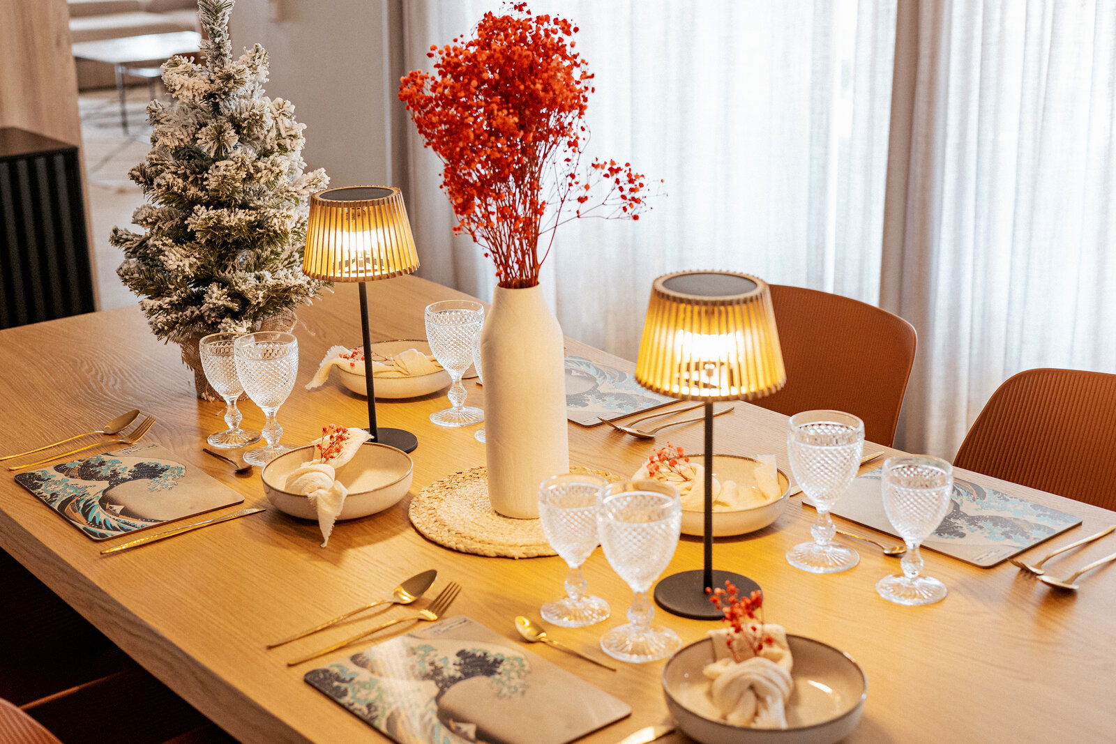 Festlich gedeckter Holzesstisch in schräger Seitenansicht mit zwei warm leuchtenden Tischlampen, Kristallgläsern, goldfarbenem Besteck, Tellern und Servietten, mittiger weißer Vase mit roten Trockenblumen sowie kleinem beschneiten Deko-Baum am Fenster.