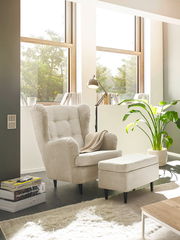 Hocker in einem modernen Wohnzimmer, seitliche Perspektive, neben einem Sessel und einer Stehlampe, mit großen Fenstern im Hintergrund.