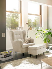 Hocker in einem modernen Wohnzimmer, seitliche Perspektive, neben einem Sessel und einer Stehlampe, mit großen Fenstern im Hintergrund.