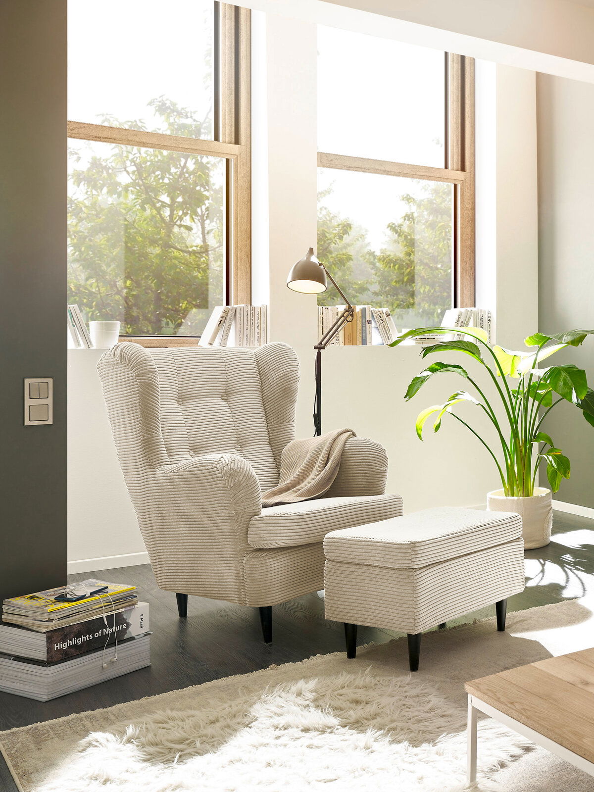 Hocker in einem modernen Wohnzimmer, seitliche Perspektive, neben einem Sessel und einer Stehlampe, mit großen Fenstern im Hintergrund.