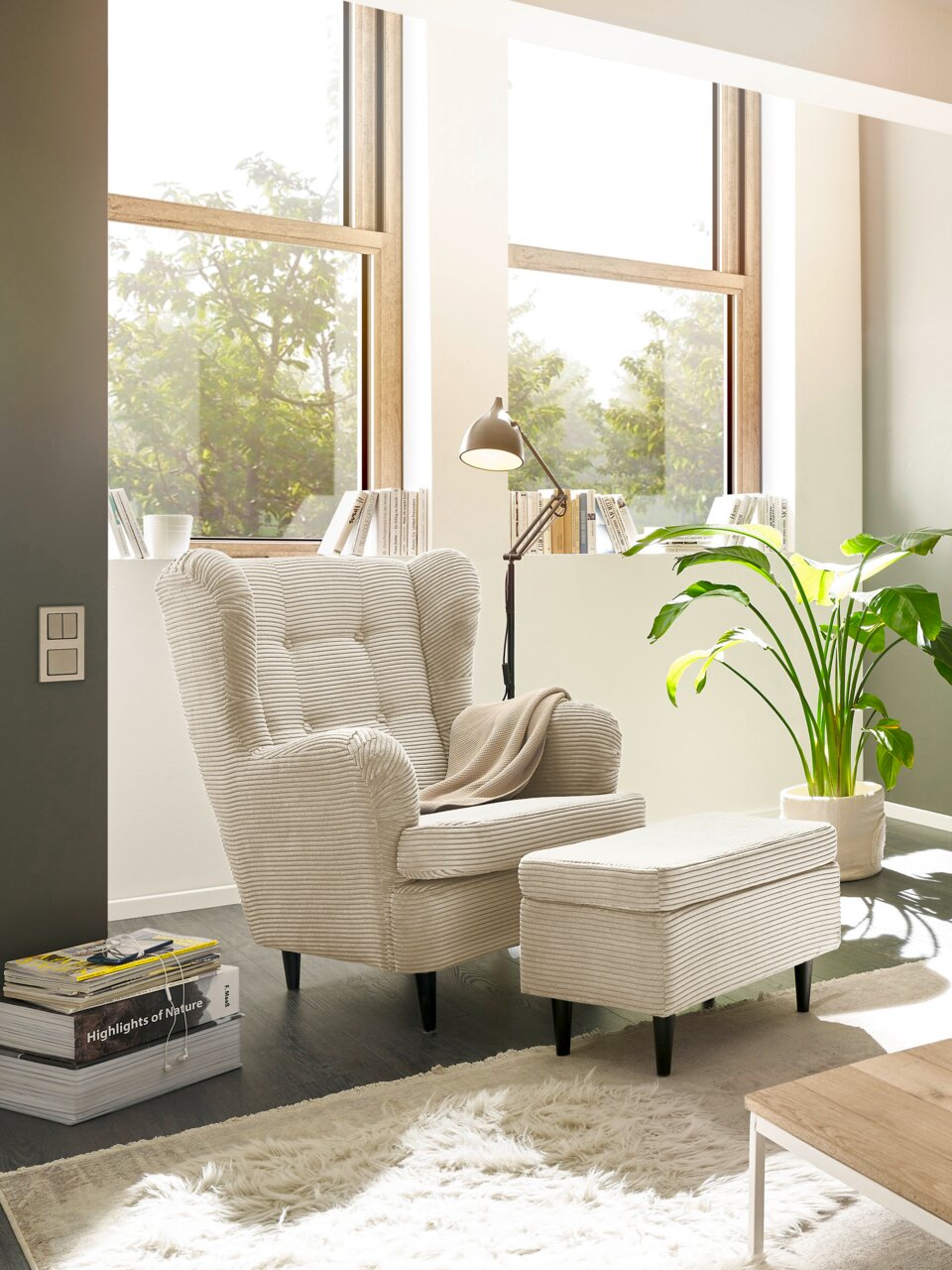 Z2 Hocker OSKAR Hocker in einem modernen Wohnzimmer, seitliche Perspektive, neben einem Sessel und einer Stehlampe, mit großen Fenstern im Hintergrund.