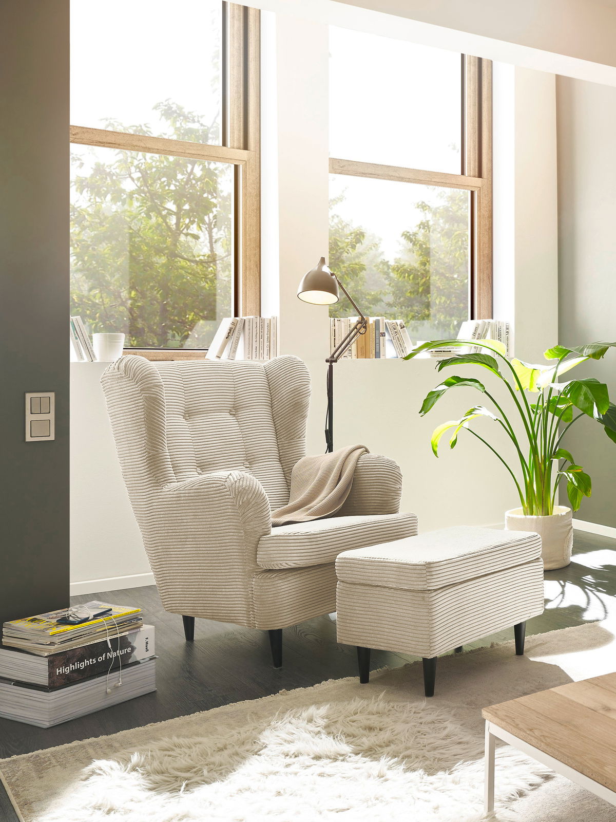 Hocker in einem modernen Wohnzimmer, seitliche Perspektive, neben einem Sessel und einer Stehlampe, mit großen Fenstern im Hintergrund.