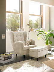 Een poef in een moderne woonkamer, gezien vanuit de zijkant, naast een fauteuil en een staande lamp, met grote ramen op de achtergrond.