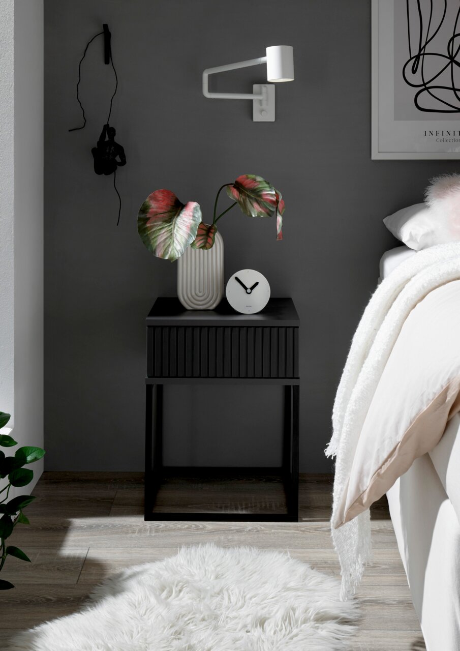 Moderne schwarze Nachtkommode mit geriffelter Front, dekoriert mit einer Vase und einer Uhr, seitliche Perspektive.