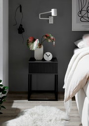 Moderne schwarze Nachtkommode mit geriffelter Front, dekoriert mit einer Vase und einer Uhr, seitliche Perspektive.