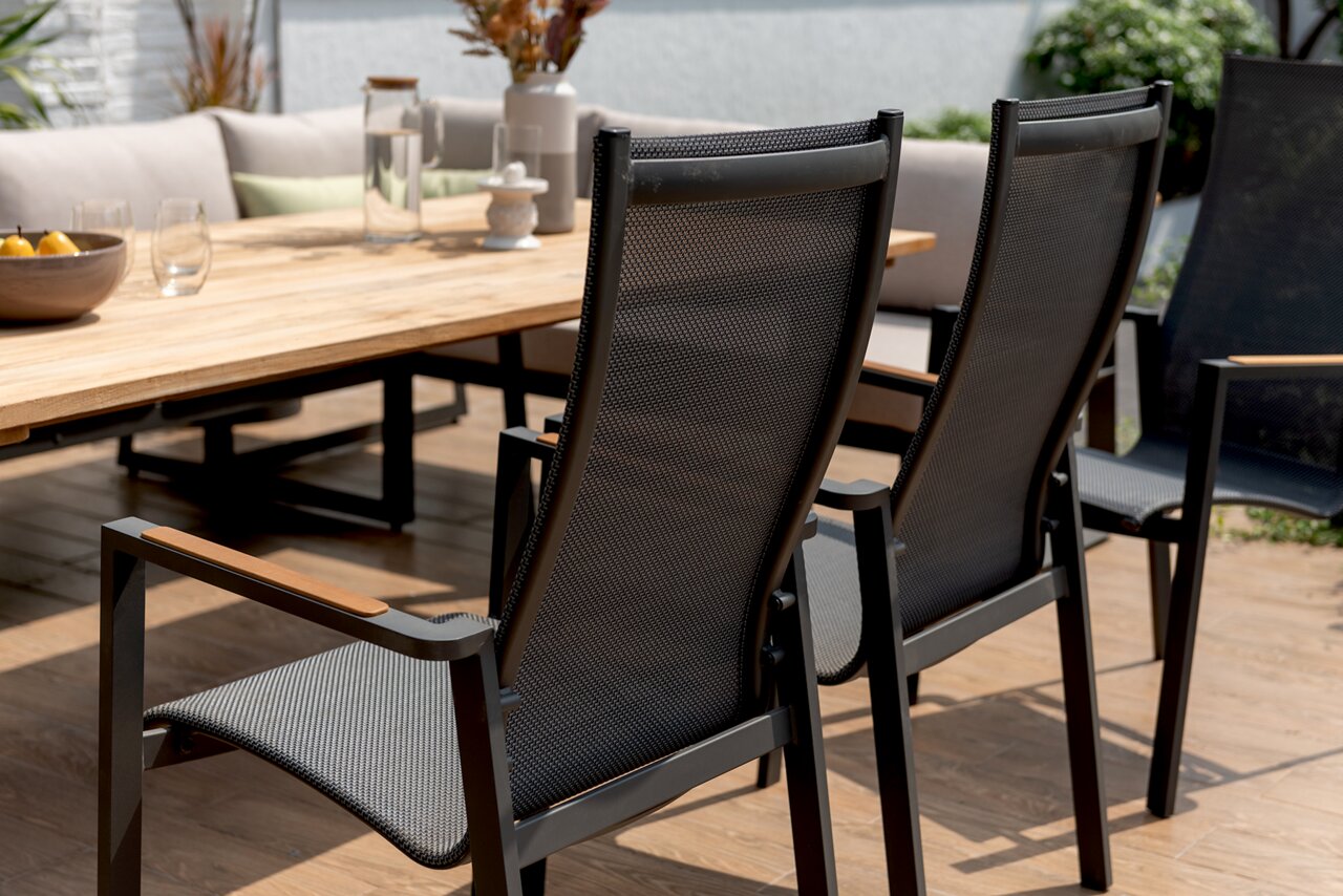 Rückansicht von zwei modernen Stapelsesseln mit schwarzem Netzgewebe und Armlehnen aus Holz, auf einer Terrasse neben einem Holztisch.