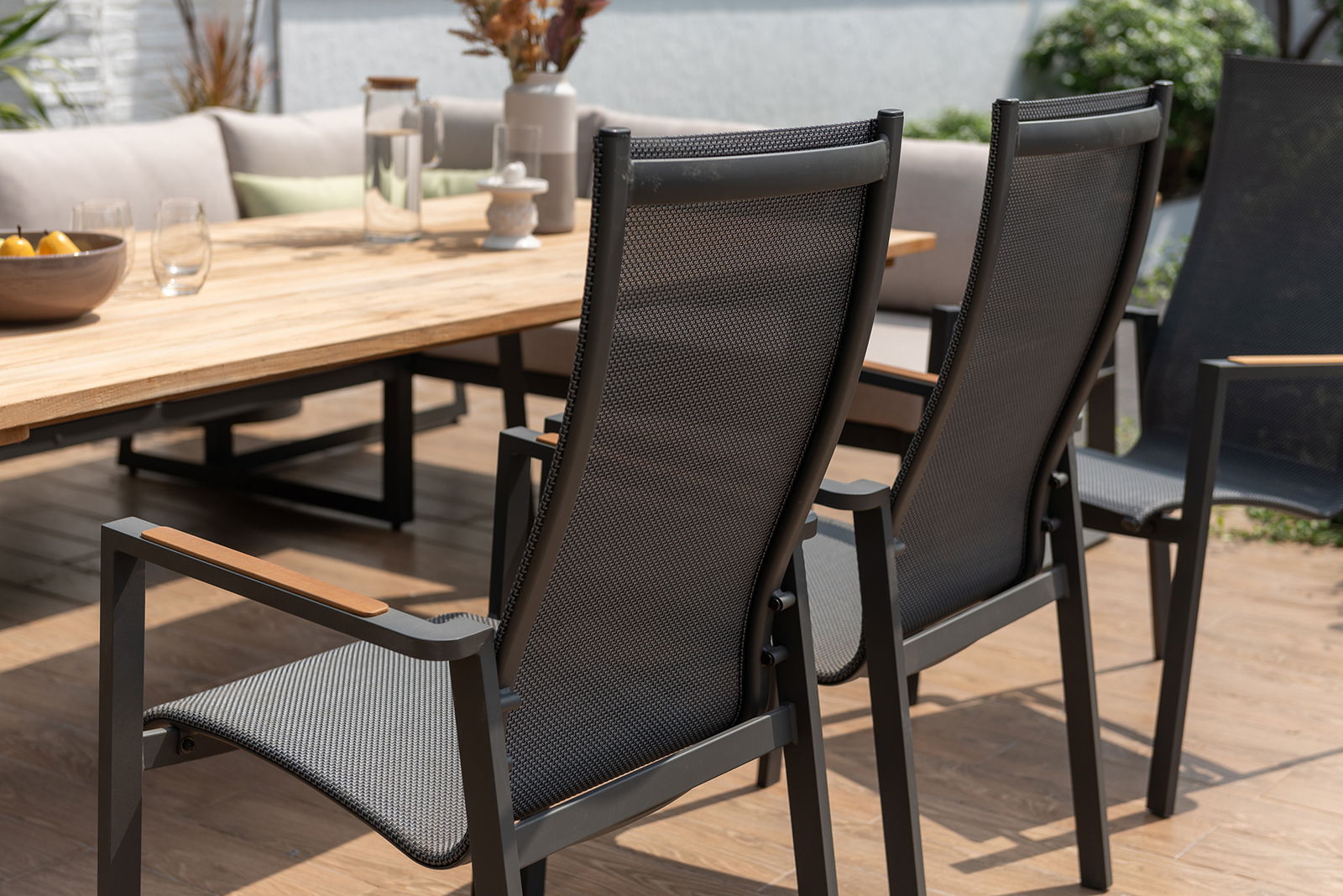 Rückansicht von zwei modernen Stapelsesseln mit schwarzem Netzgewebe und Armlehnen aus Holz, auf einer Terrasse neben einem Holztisch.