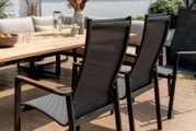 Achterkant van twee moderne stapelstoelen met zwart gaas en houten armleuningen, op een terras naast een houten tafel.