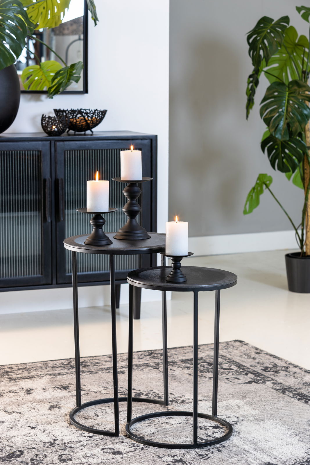 Zwei schwarze Beistelltische mit runden Tischplatten, dekoriert mit Kerzenhaltern und brennenden Kerzen, in einem modernen Wohnzimmer mit Pflanzen und einem Schrank im Hintergrund. Perspektive von vorne.