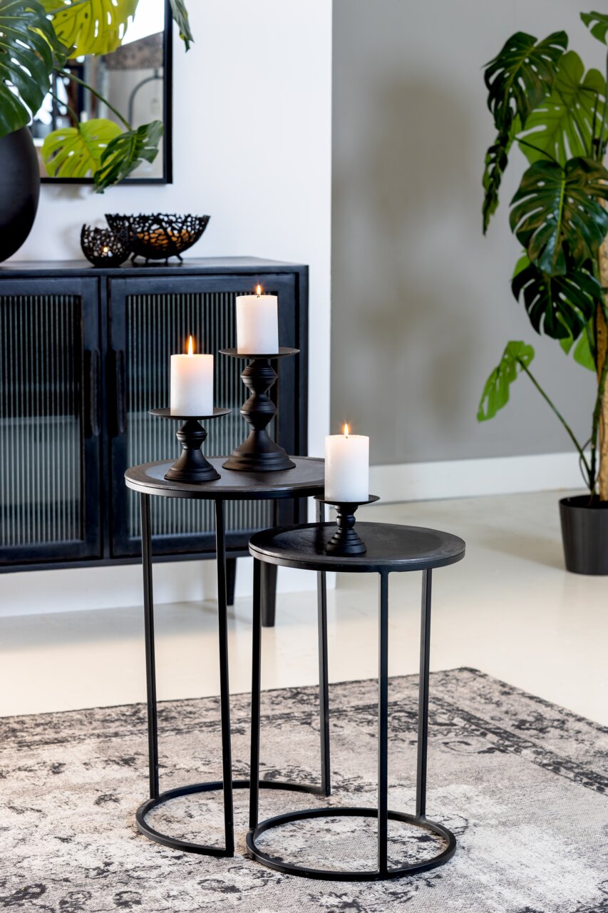 Zwei schwarze Beistelltische mit runden Tischplatten, dekoriert mit Kerzenhaltern und brennenden Kerzen, in einem modernen Wohnzimmer mit Pflanzen und einem Schrank im Hintergrund. Perspektive von vorne.