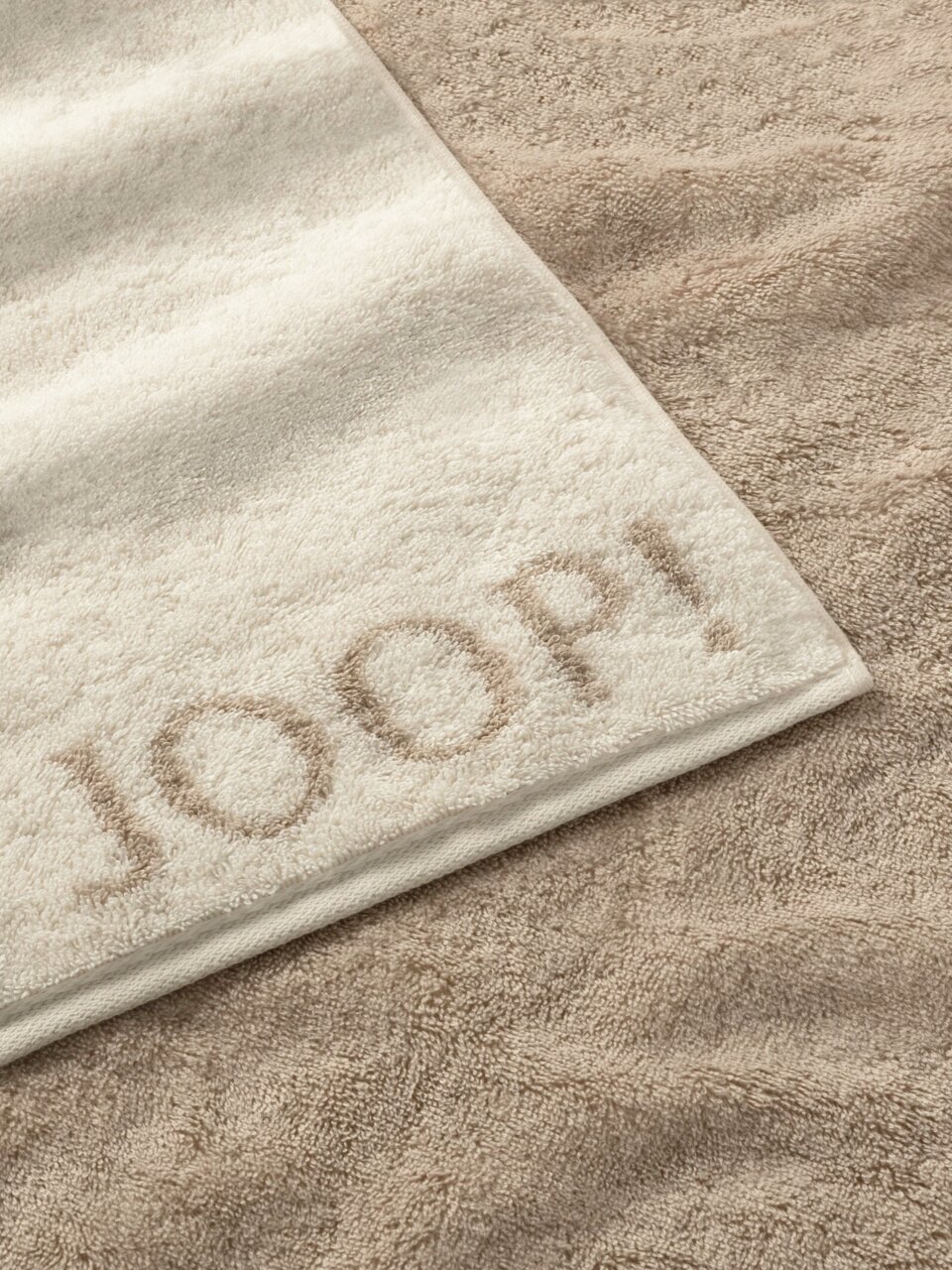 Nahaufnahme eines beigen Duschtuchs mit eingewebtem Schriftzug 'JOOP!' auf einem sandfarbenen Hintergrund.