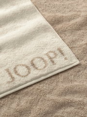 Nahaufnahme eines beigen Duschtuchs mit eingewebtem Schriftzug 'JOOP!' auf einem sandfarbenen Hintergrund.