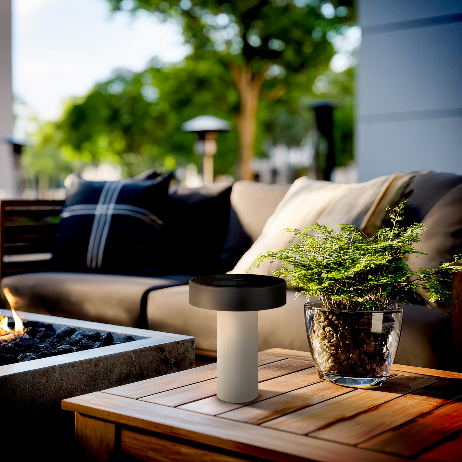 Moderne kabellose Outdoor-Tischlampe mit schwarzem, flachem Lampenkopf und geriffeltem weißem Sockel auf einem Holztisch neben einer Pflanze und Feuerstelle, fotografiert aus leicht erhöhter Frontperspektive auf einer Lounge-Terrasse.