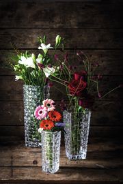 Drei elegante, klare Glasvasen mit Blumenarrangements, fotografiert aus einer leicht erhöhten Perspektive vor einem rustikalen Holzhintergrund.