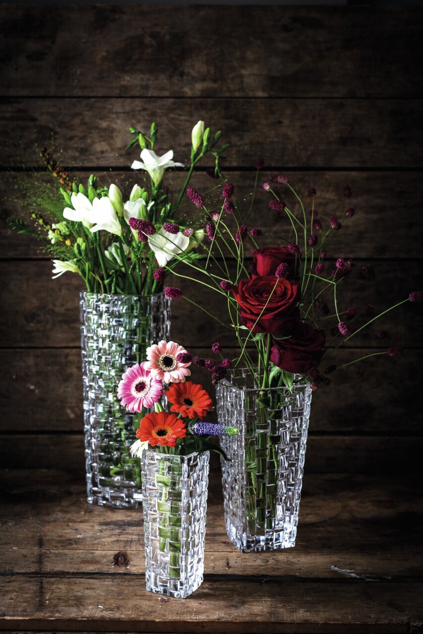 Drei elegante, klare Glasvasen mit Blumenarrangements, fotografiert aus einer leicht erhöhten Perspektive vor einem rustikalen Holzhintergrund.