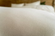 Nahaufnahme einer strukturierten weißen Bettwäsche mit Fokus auf das Gewebe, im Hintergrund unscharf ein Bett zu sehen.