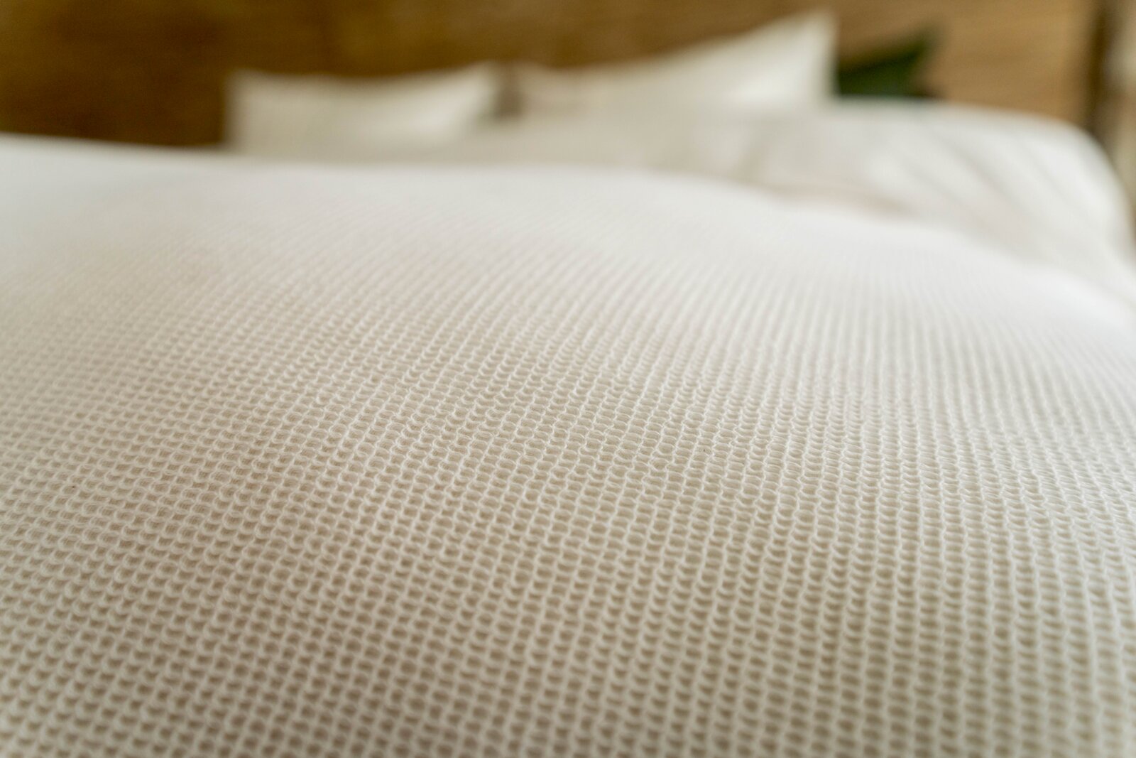 Nahaufnahme einer strukturierten weißen Bettwäsche mit Fokus auf das Gewebe, im Hintergrund unscharf ein Bett zu sehen.