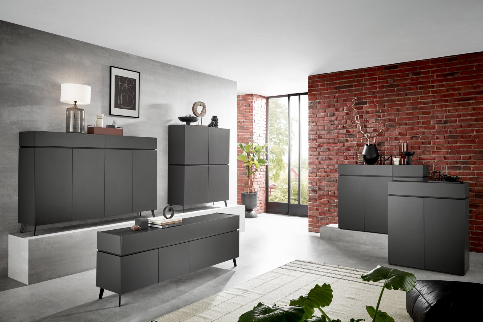 Voleo Kommode UNIQUE Moderne, dunkelgraue Kommode in einem stilvollen Wohnzimmer mit Ziegelwand und großen Fenstern, seitliche Perspektive.