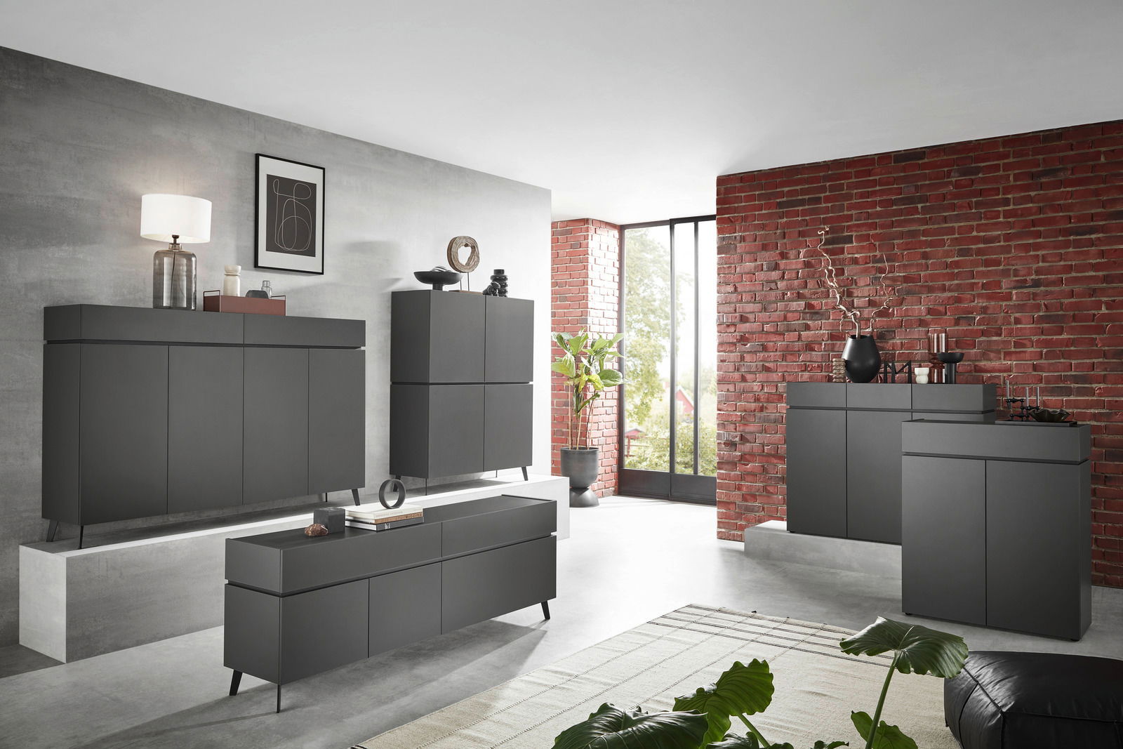 Moderne, dunkelgraue Kommode in einem stilvollen Wohnzimmer mit Ziegelwand und großen Fenstern, seitliche Perspektive.