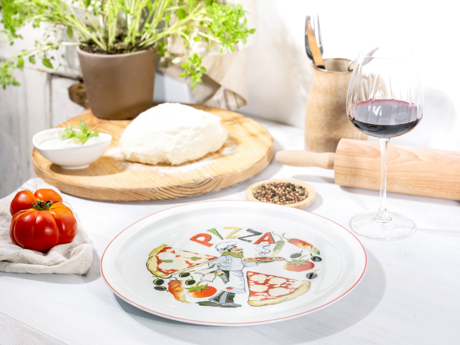 Weißer Pizzateller mit farbigem Pizza-Motiv und Schriftzug „PIZZA“ auf gedecktem Tisch, mit Teig, Tomaten und Rotweinglas im Hintergrund, aufgenommen in leicht schräger Frontansicht von oben.