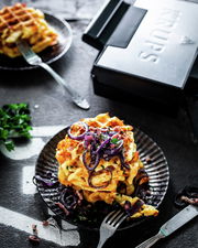 Nahaufnahme eines Waffeleisens neben einem Teller mit frisch gebackenen Waffeln, garniert mit roten Zwiebeln und Kräutern.