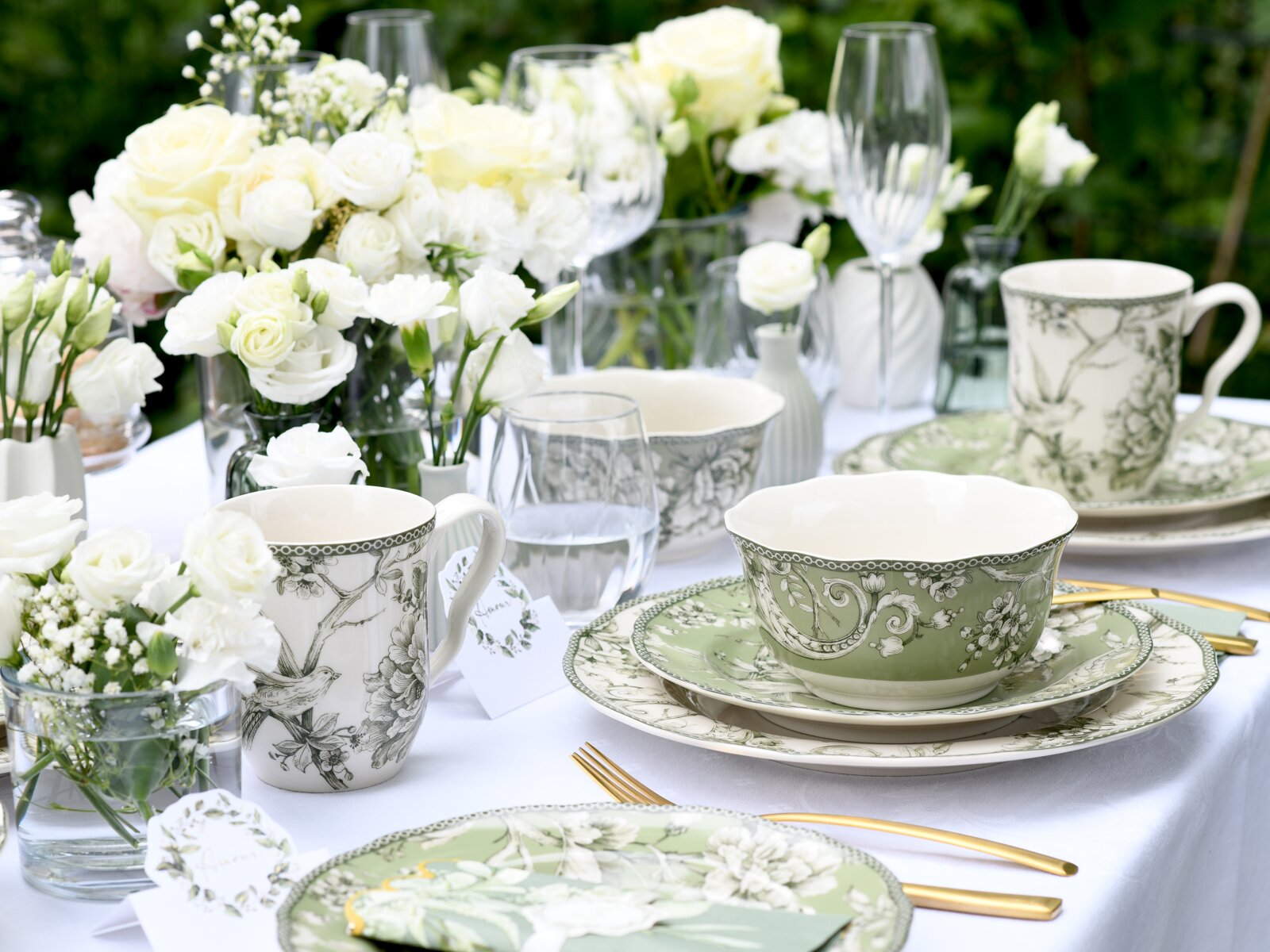 Elegantes Outdoor-Tischgedeck mit grün-weißem Porzellan (Schale, Teller und Tasse mit floralen Mustern), goldfarbenem Besteck, Glaswaren und weißen Rosen; Perspektive: leicht erhöht aus schräger Frontansicht.