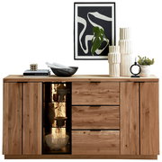 Frontansicht einer modernen Holz-Kommode/Sideboard in Eiche-Optik mit zwei Türen, drei Schubladen und beleuchtetem Glasvitrinenfach, dekoriert mit Vasen, Büchern und Pflanzen; darüber abstraktes Wandbild