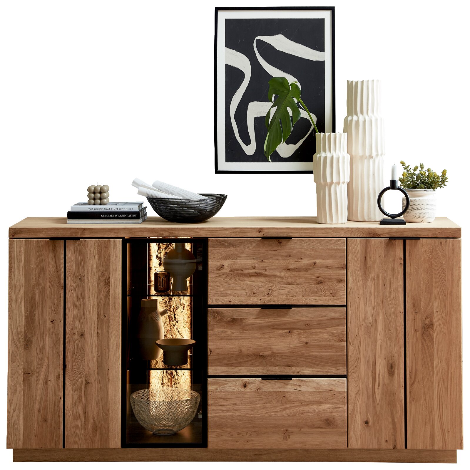 Cantus Sideboard DONNA Frontansicht einer modernen Holz-Kommode/Sideboard in Eiche-Optik mit zwei Türen, drei Schubladen und beleuchtetem Glasvitrinenfach, dekoriert mit Vasen, Büchern und Pflanzen; darüber abstraktes Wandbild