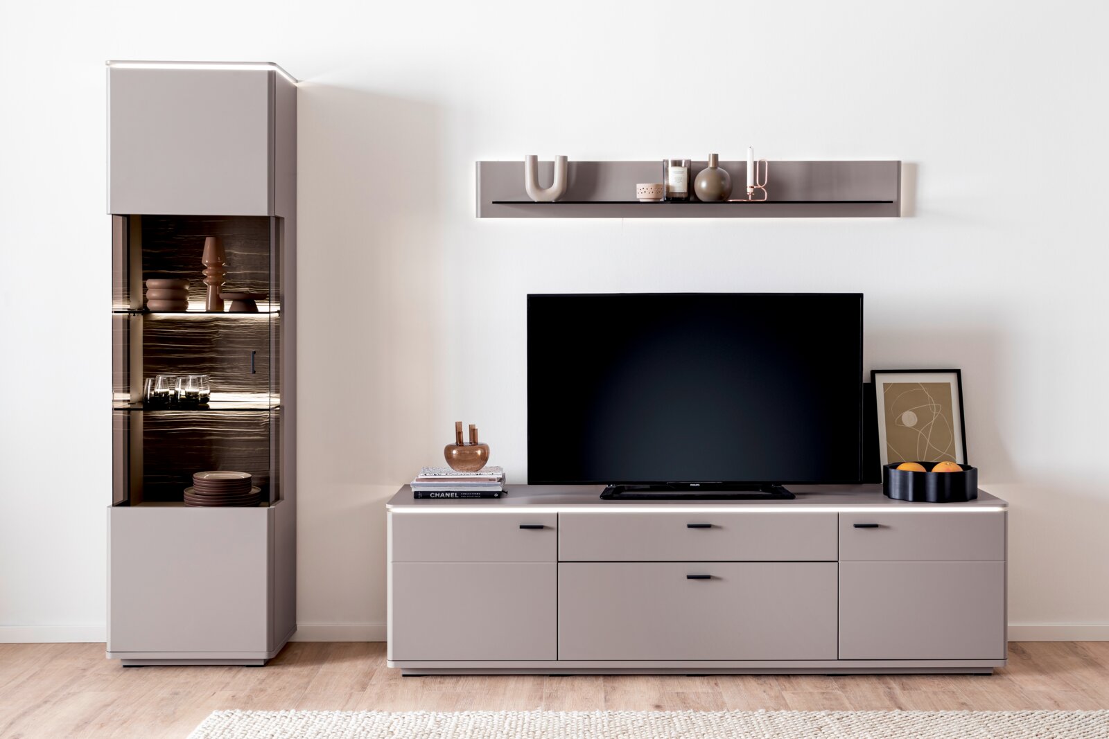 Frontansicht einer modernen Wohnwand in Hellgrau mit Lowboard und Hängeregal, links eine Vitrine mit Glasfront und Holzoptik, in der Mitte ein großer Fernseher auf dem Lowboard, dekoriert mit Vasen, Büchern und Schale.
