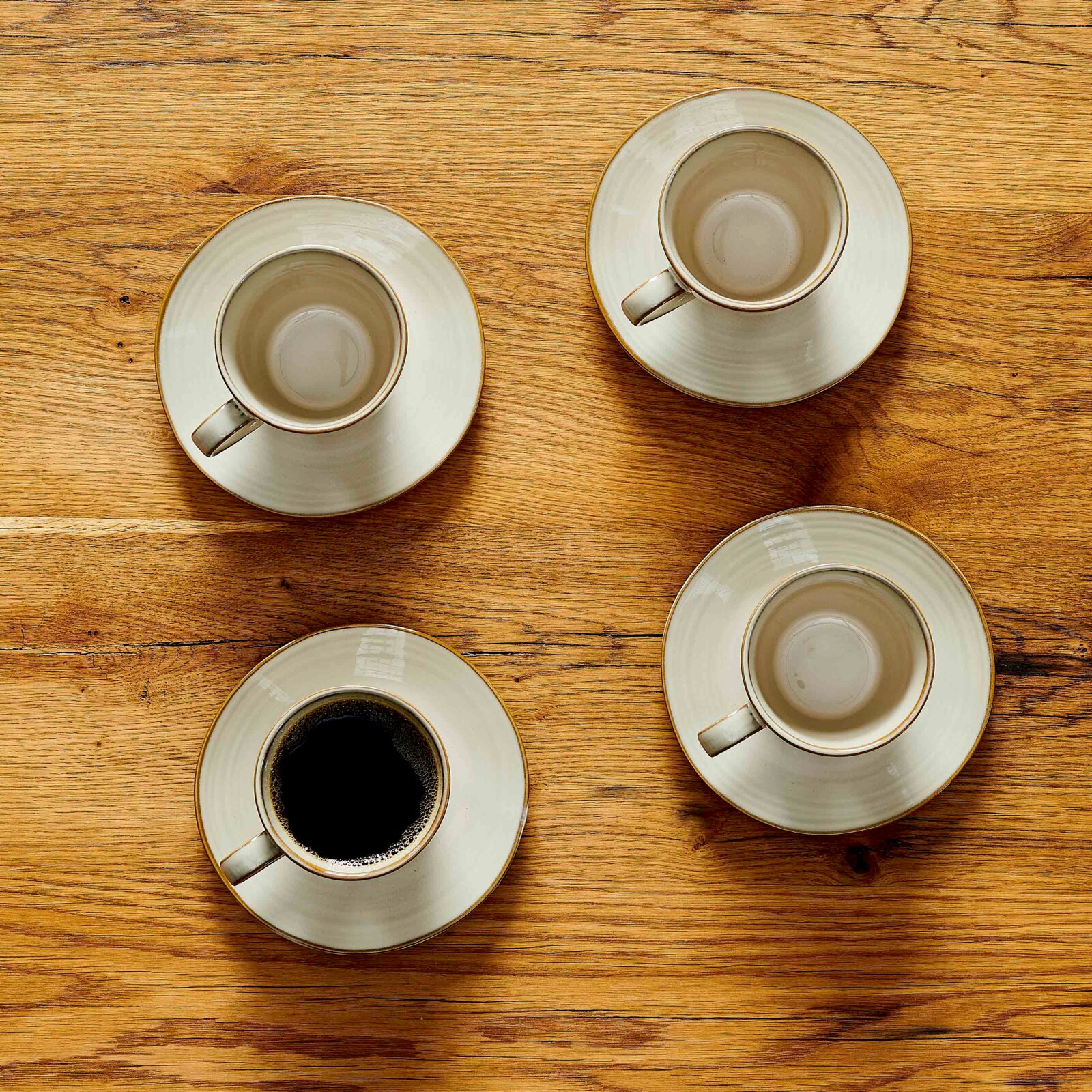 Draufsicht auf mehrere cremefarbene Kaffeetassen mit Untertassen auf einem rustikalen Holztisch, eine Tasse mit schwarzem Kaffee gefüllt