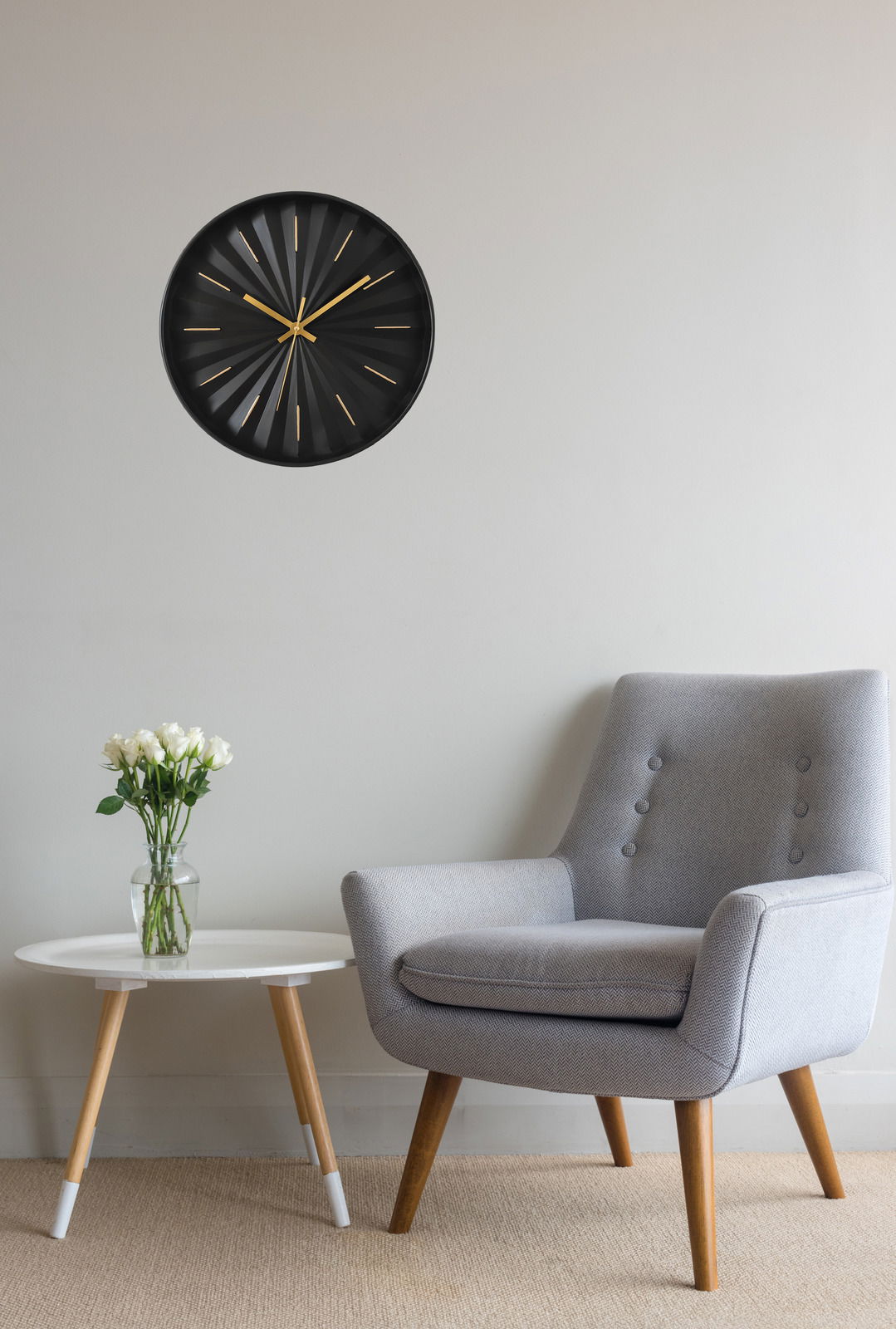Moderne schwarze Wanduhr mit goldenen Zeigern und Markierungen, an einer hellen Wand über einem grauen Sessel und einem weißen Tisch mit Blumen, aus frontaler Perspektive.