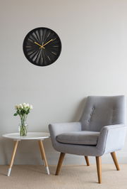 Moderne schwarze Wanduhr mit goldenen Zeigern und Markierungen, an einer hellen Wand über einem grauen Sessel und einem weißen Tisch mit Blumen, aus frontaler Perspektive.