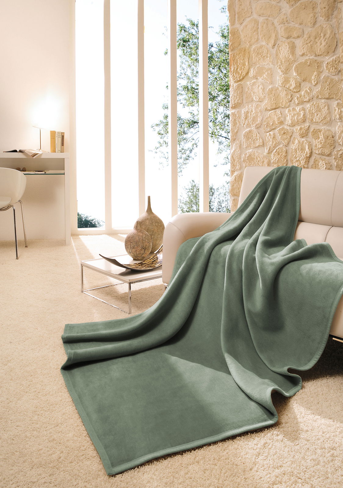 Grüne Wohndecke 150 x 200 cm auf einem Sofa in einem modernen Wohnzimmer mit Steinwand und großen Fenstern, seitliche Perspektive