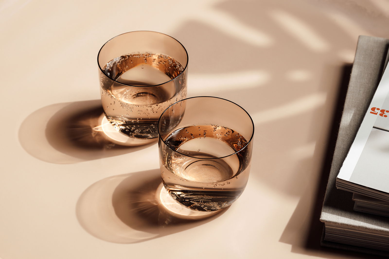 Like by Villeroy & Boch Wasserglas Set 2-tlg. LIKE CLAY Zwei elegante Wassergläser mit sprudelndem Wasser, von oben fotografiert, auf einem Tisch neben einem Stapel Bücher.