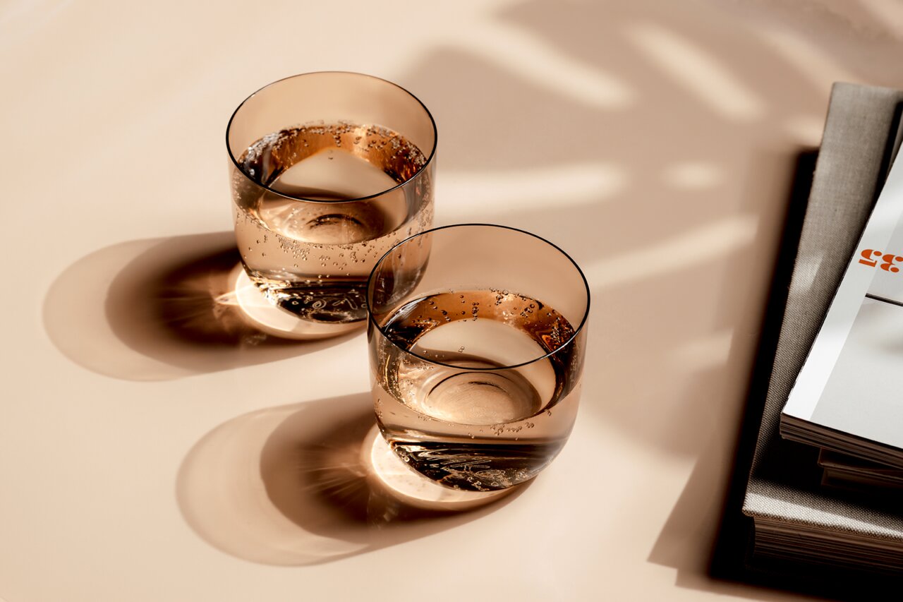 Like by Villeroy & Boch Wasserglas Set 2-tlg. LIKE CLAY Zwei elegante Wassergläser mit sprudelndem Wasser, von oben fotografiert, auf einem Tisch neben einem Stapel Bücher.