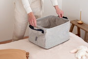 Faltbarer grauer Wäschekorb mit schwarzen Griffen, auf einem Bett platziert, von einer Person gehalten, seitliche Perspektive.