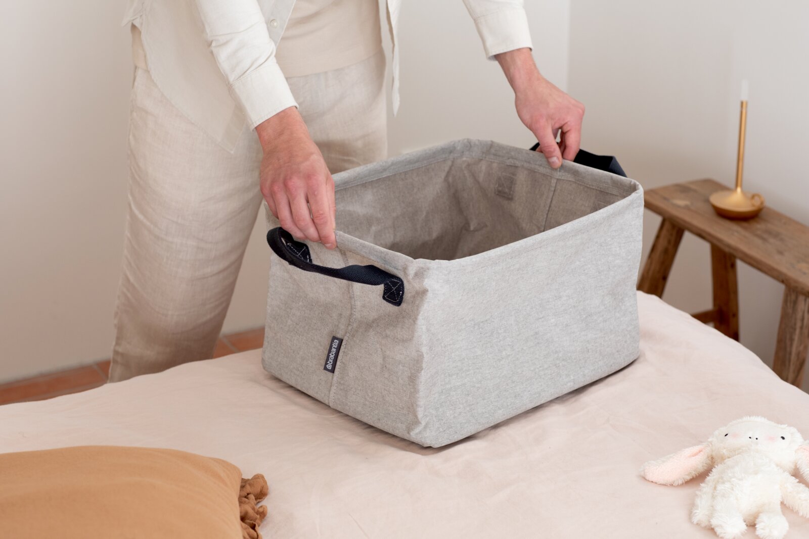 Brabantia Faltbarer Wäschekorb MINDSET Faltbarer grauer Wäschekorb mit schwarzen Griffen, auf einem Bett platziert, von einer Person gehalten, seitliche Perspektive.