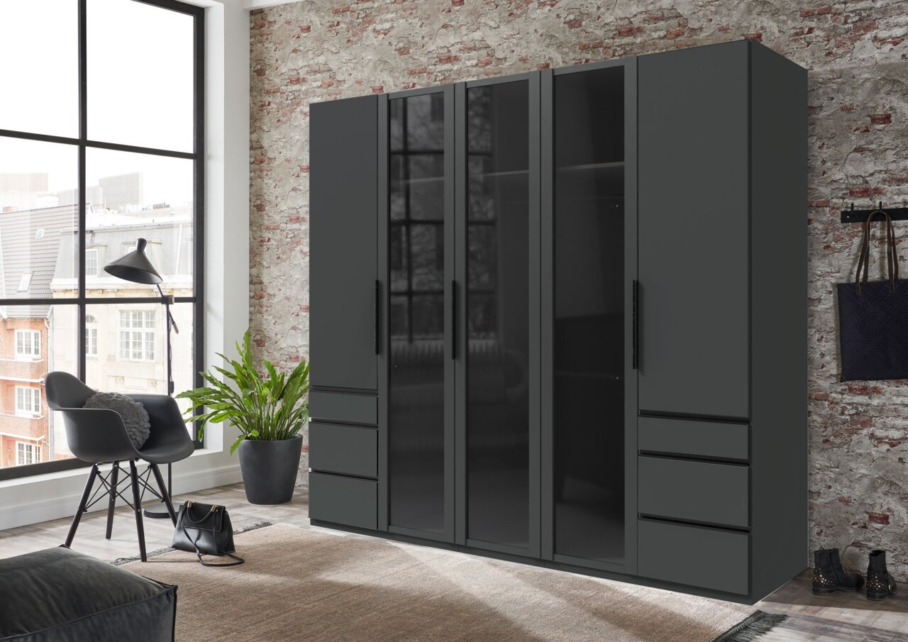 Moderner 5-türiger Drehtürenschrank in dunklem Design, frontal abgebildet in einem stilvollen Raum mit Backsteinwand und großen Fenstern.
